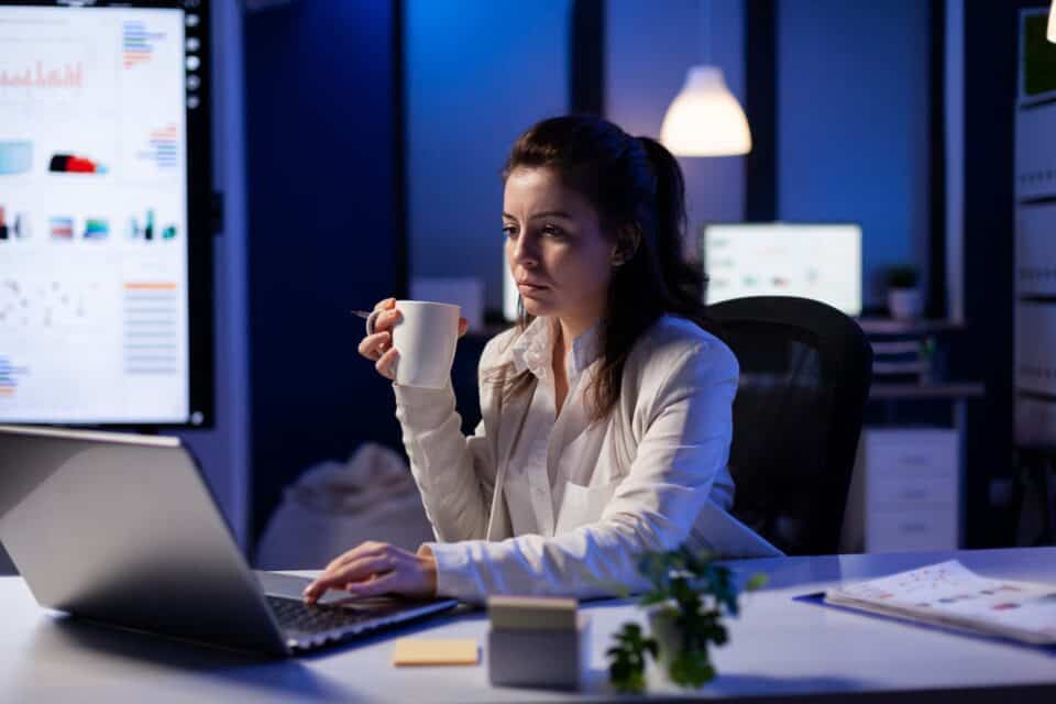 A tired woman working on a laptop at night and holding a cup of coffee.